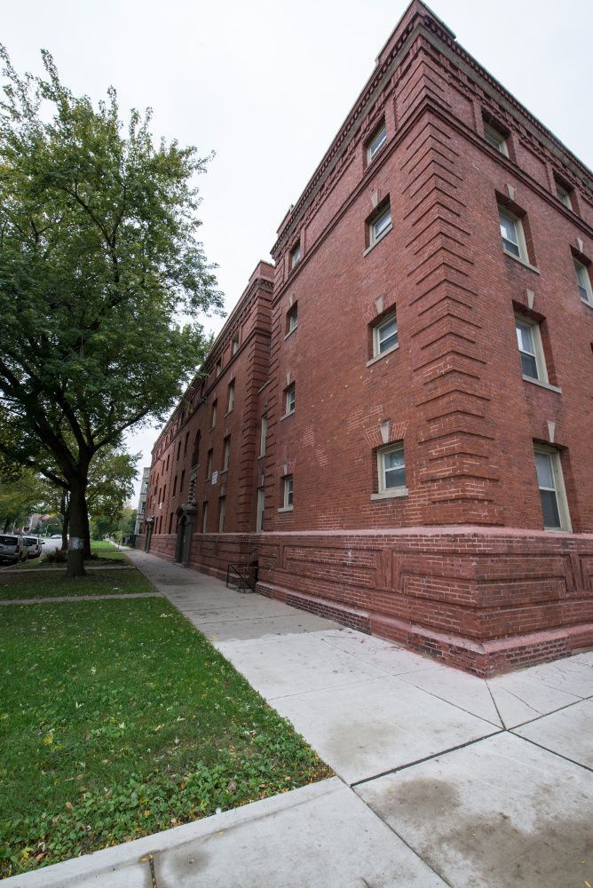 Brick apartment building corner with sidewalk, grass, and tree on an overcast day.