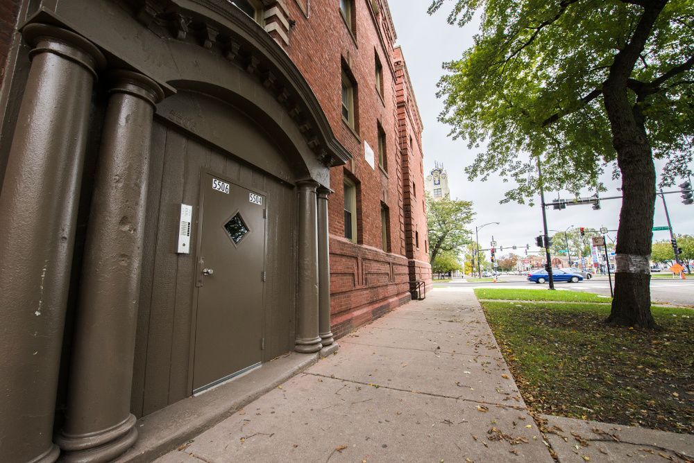 Brown doorway with columns, red brick building, sidewalk, street, and a tree.