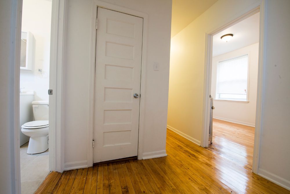 Hallway with wooden floor, white walls and doors. Bathroom on left, room with window on right.