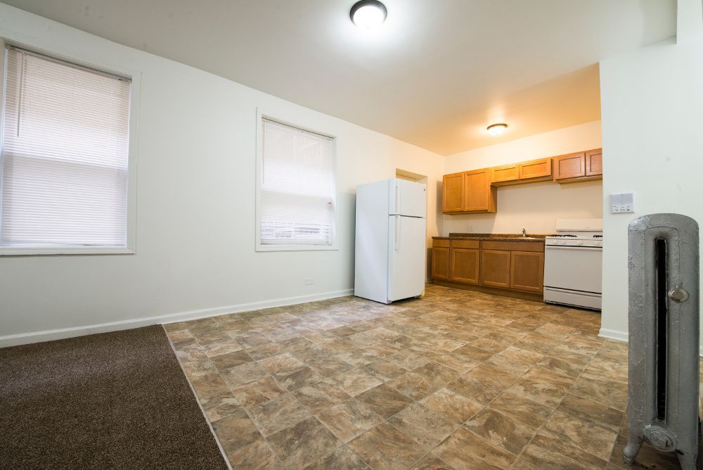 Empty apartment interior with kitchen, windows, and radiator. Brown and tan floor and beige walls.