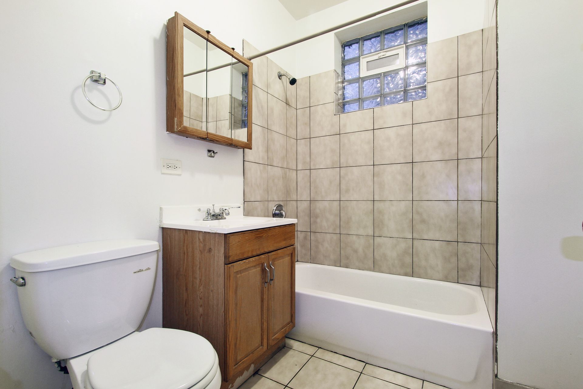 Small bathroom with toilet, sink, mirror, and bathtub/shower. Neutral color scheme, window with glass blocks.