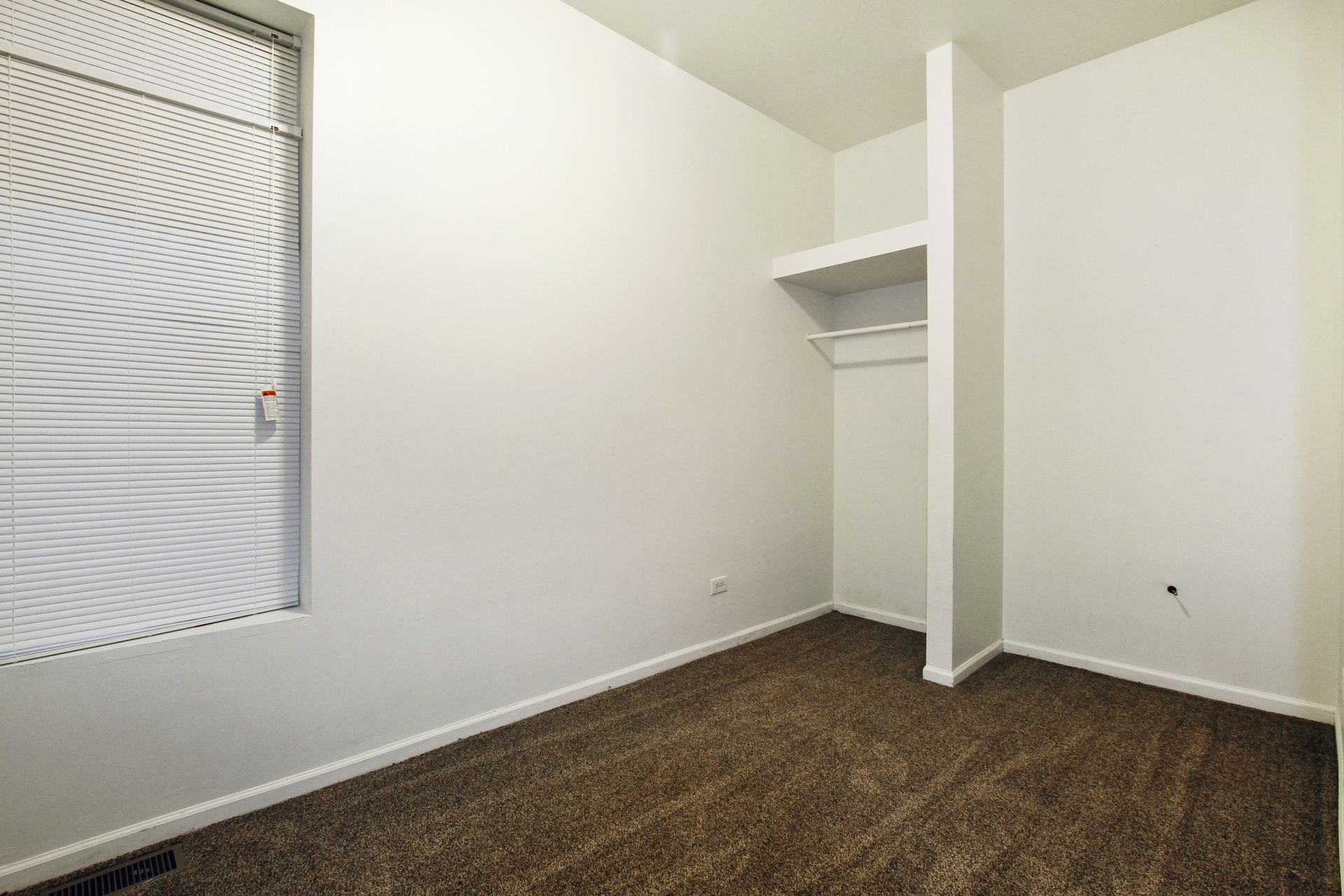 Empty room with brown carpet, white walls, closet area, and barred window.