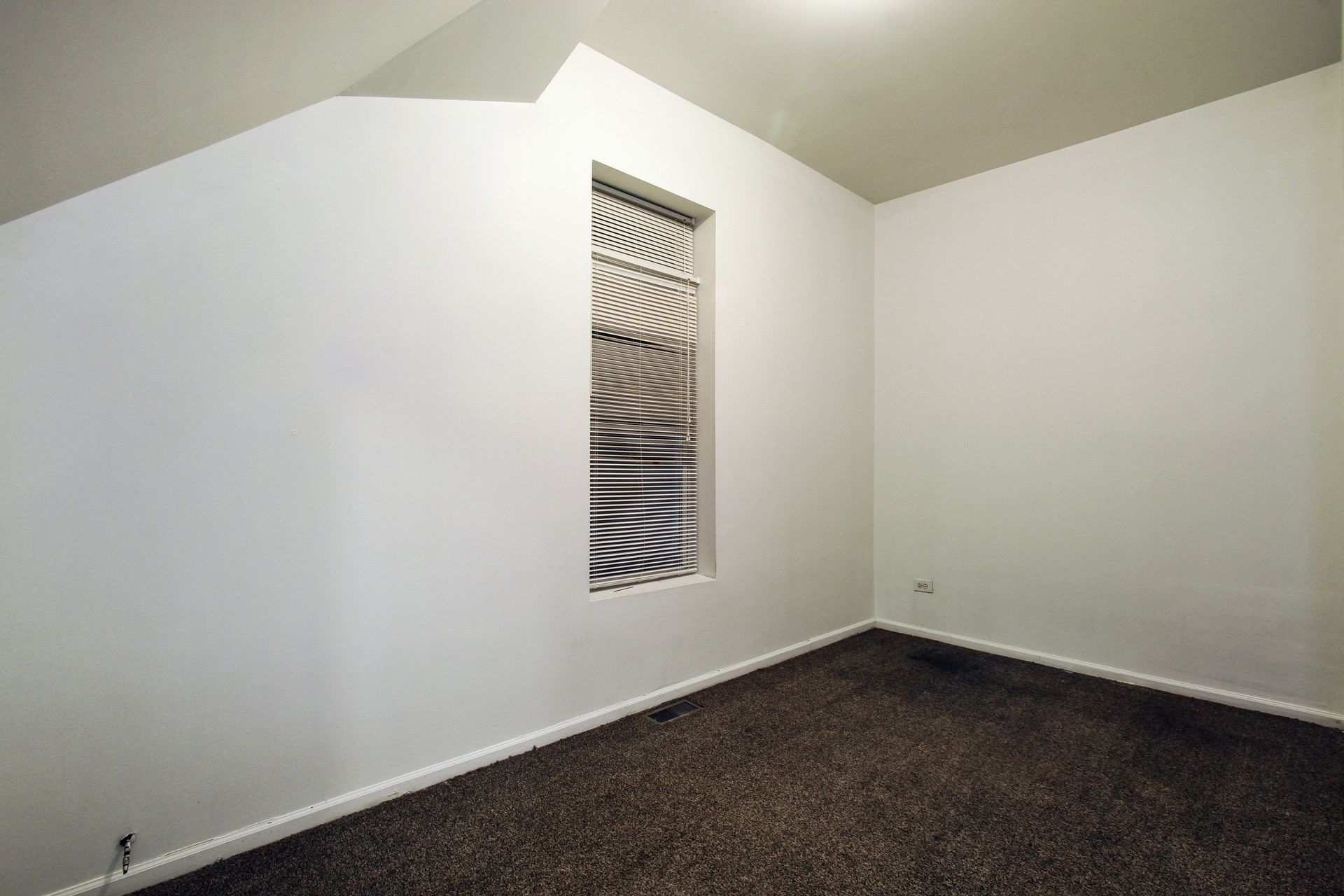 Empty room with dark brown carpet, white walls, and a narrow window with closed blinds.