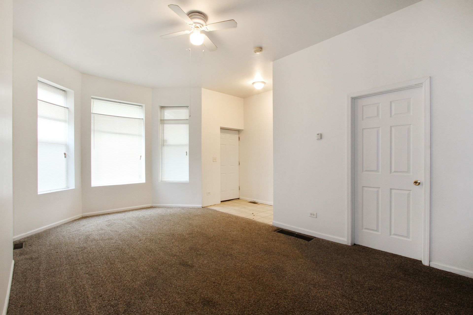 Empty room with brown carpet, white walls, three windows with blinds, and a white door.