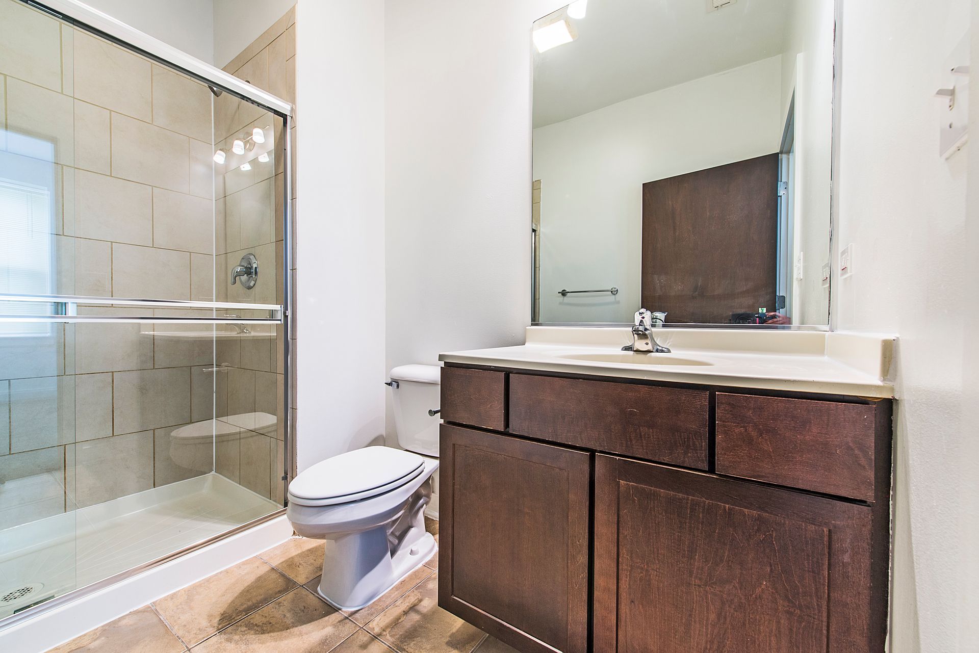Bathroom with shower, toilet, vanity, and large mirror. Light tan tile and dark brown cabinetry.