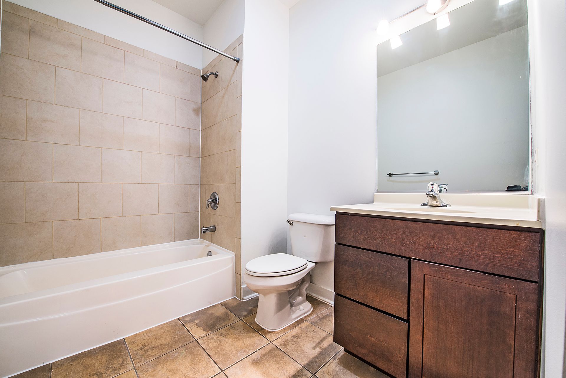 Bathroom with white bathtub, toilet, and vanity with a mirror. Tan tile on the floor and wall.