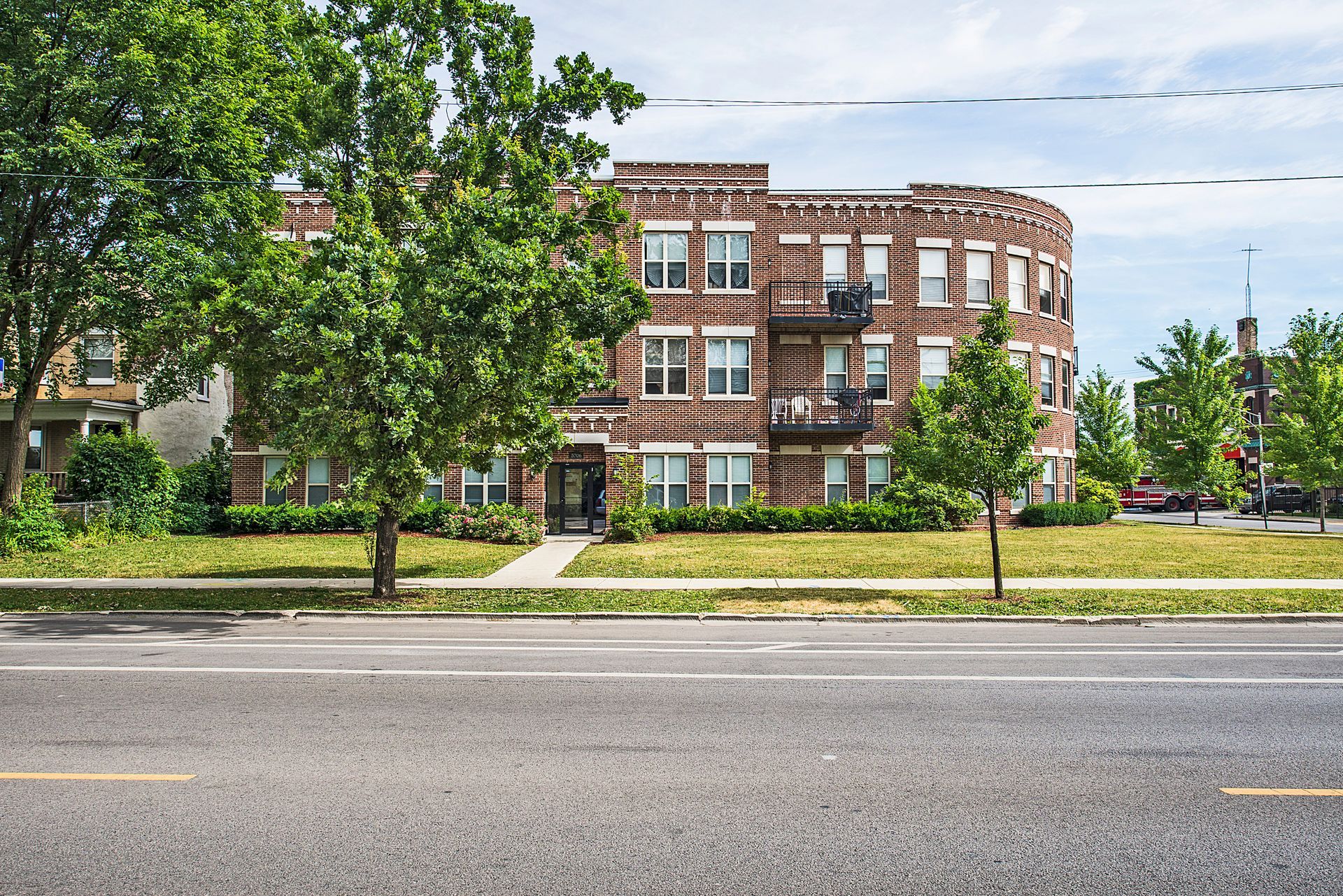 Brick apartment building with green trees, sidewalk, and street.
