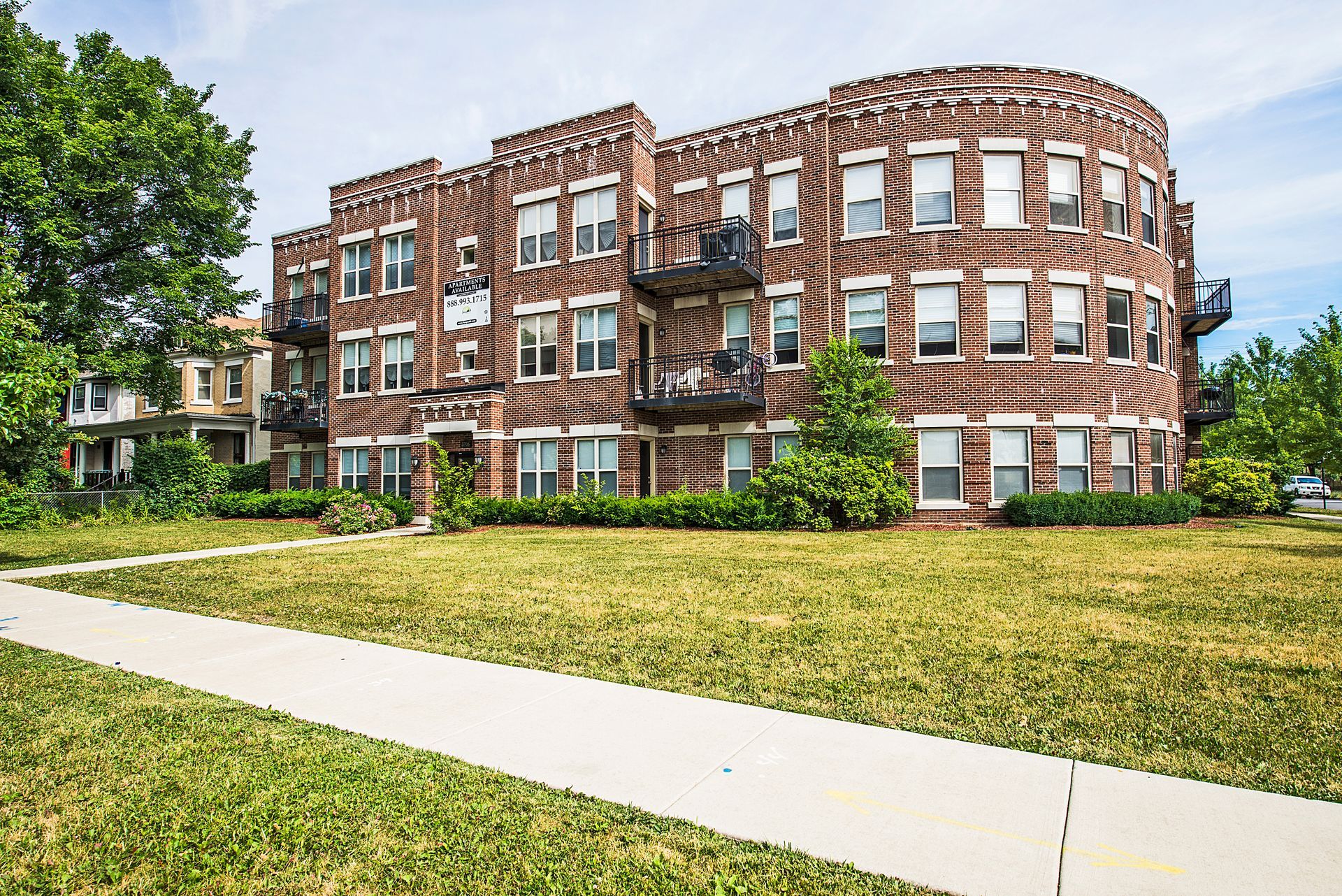 Brick apartment building with balconies, set in grassy yard, sidewalk in foreground.