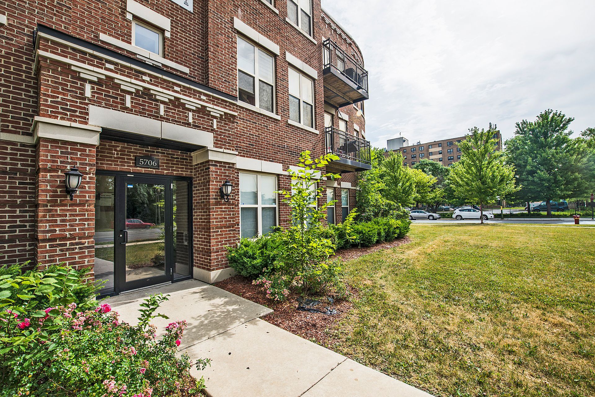 Brick apartment building entrance with glass doors, sidewalk, and landscaping.