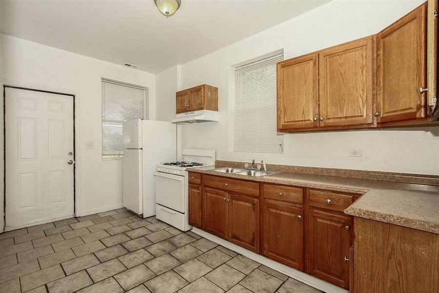 Kitchen with light-colored cabinets, white appliances, and tiled floor; door on left, windows in background.