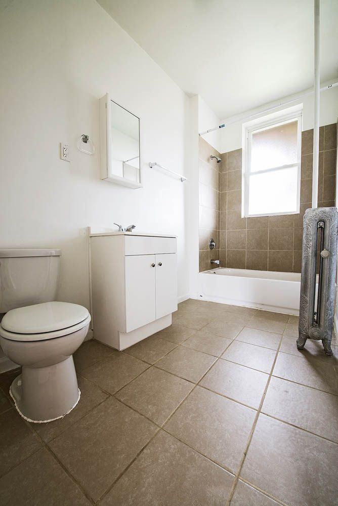Bathroom with a toilet, sink cabinet, and bathtub. Beige tiled floor and walls. Radiator visible.