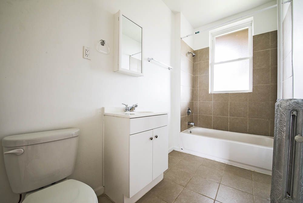 Small, white bathroom with toilet, vanity, and bathtub. Beige tiled wall and window.