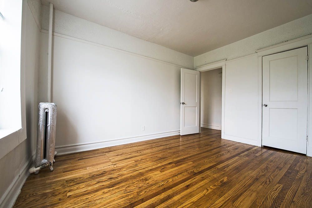 Empty room with hardwood floors, white walls, two closed doors, and a radiator.
