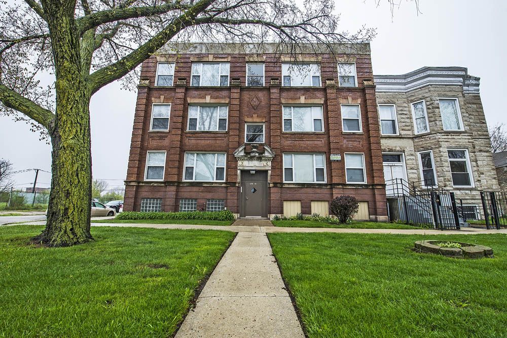 Three-story red brick apartment building with a concrete walkway and green lawn on a cloudy day.