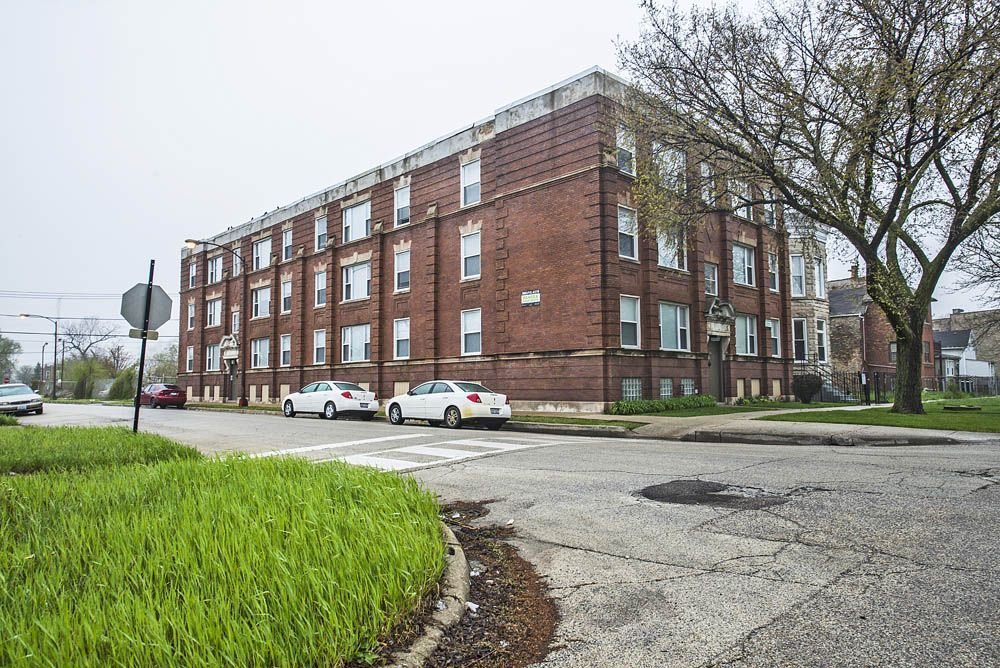 Brick apartment building on a street corner, cars parked, overcast sky.
