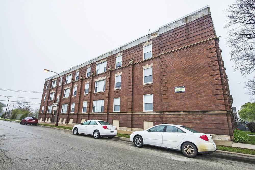 Brick apartment building on a street with parked white cars. Overcast sky.