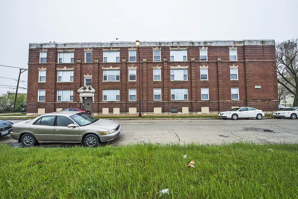 Three-story brick apartment building with cars parked on the street in front of overgrown grass.