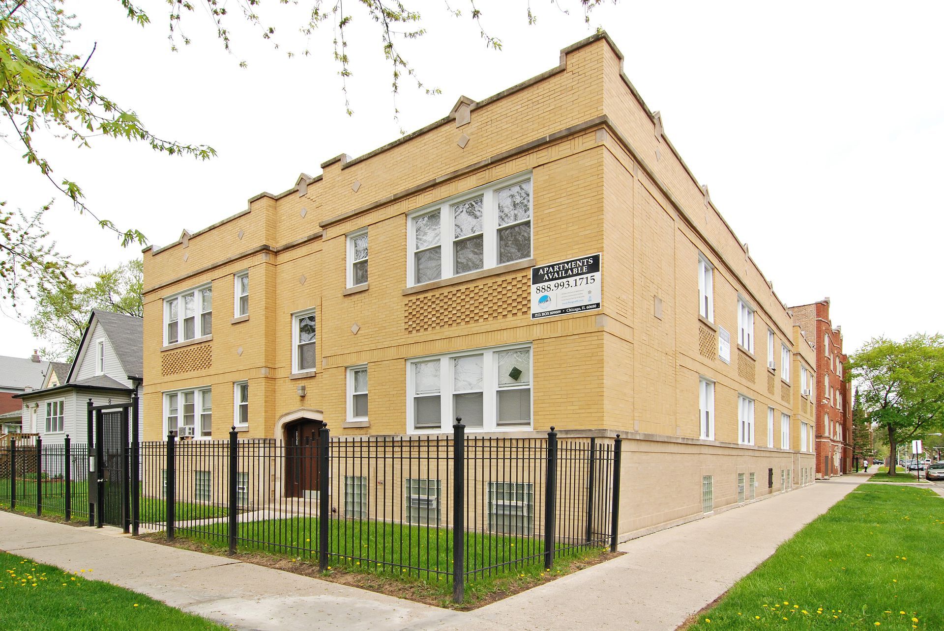 Two-story tan brick apartment building with black fence, sidewalk, and green grass.