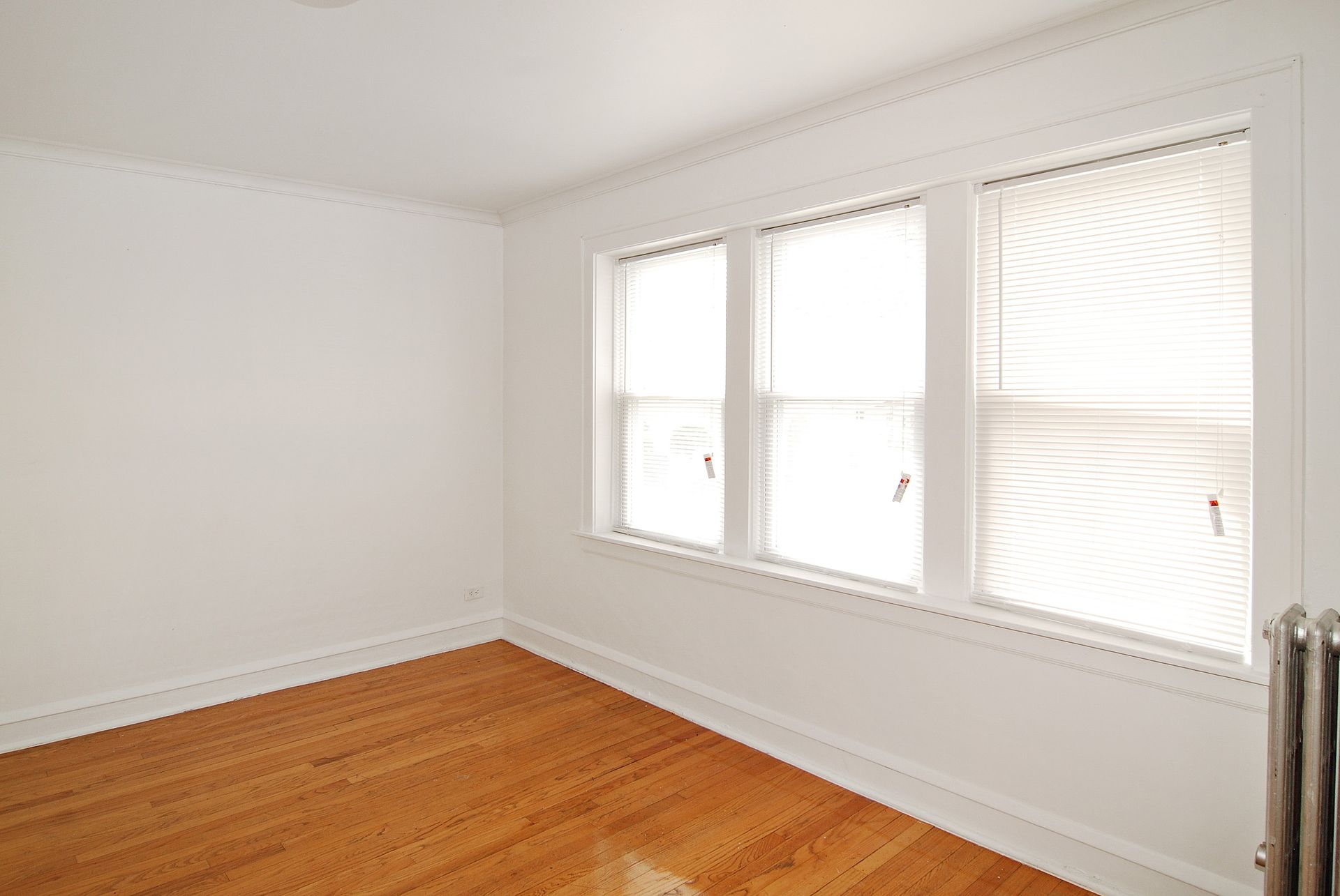 Empty room with wood floor, white walls, three windows with blinds, and radiator.
