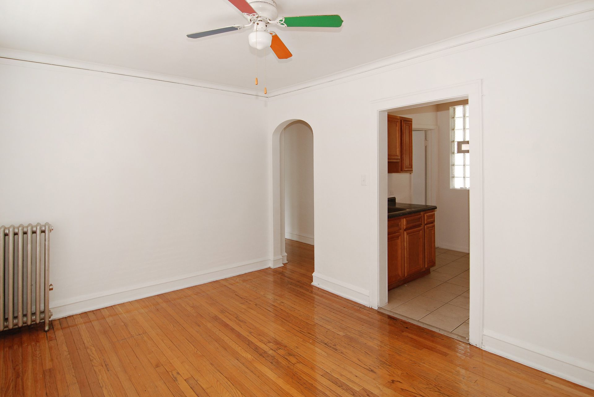 Empty room with hardwood floors, radiator, archway to kitchen with wood cabinets, and a colorful ceiling fan.