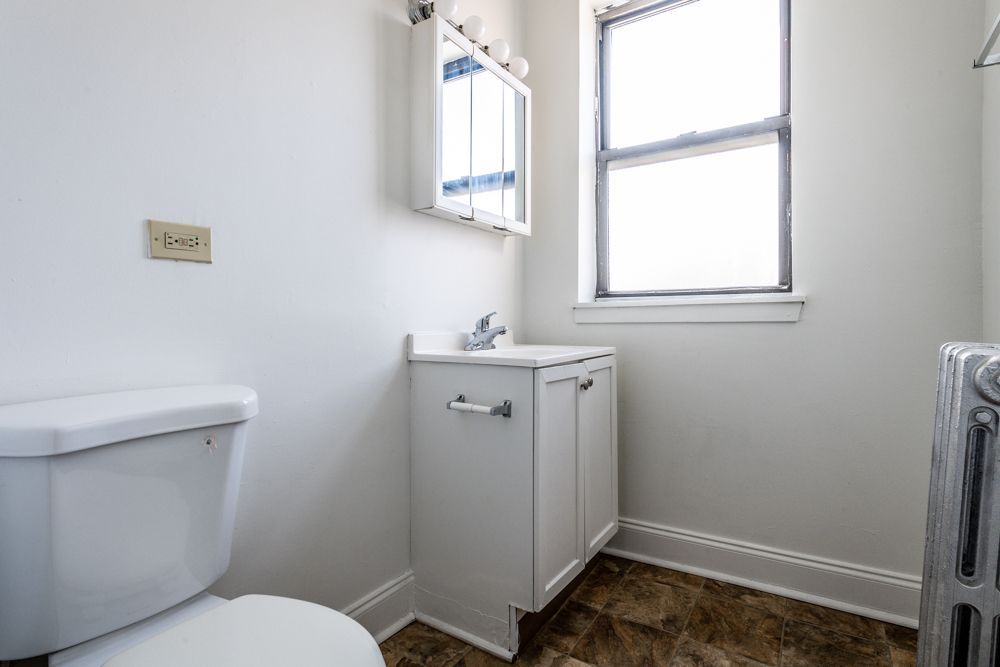 Small bathroom with a toilet, sink cabinet, mirror, window, and radiator. White walls, patterned floor.