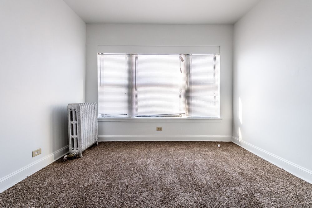 Empty room with beige carpet, white walls, window with blinds, and radiator.