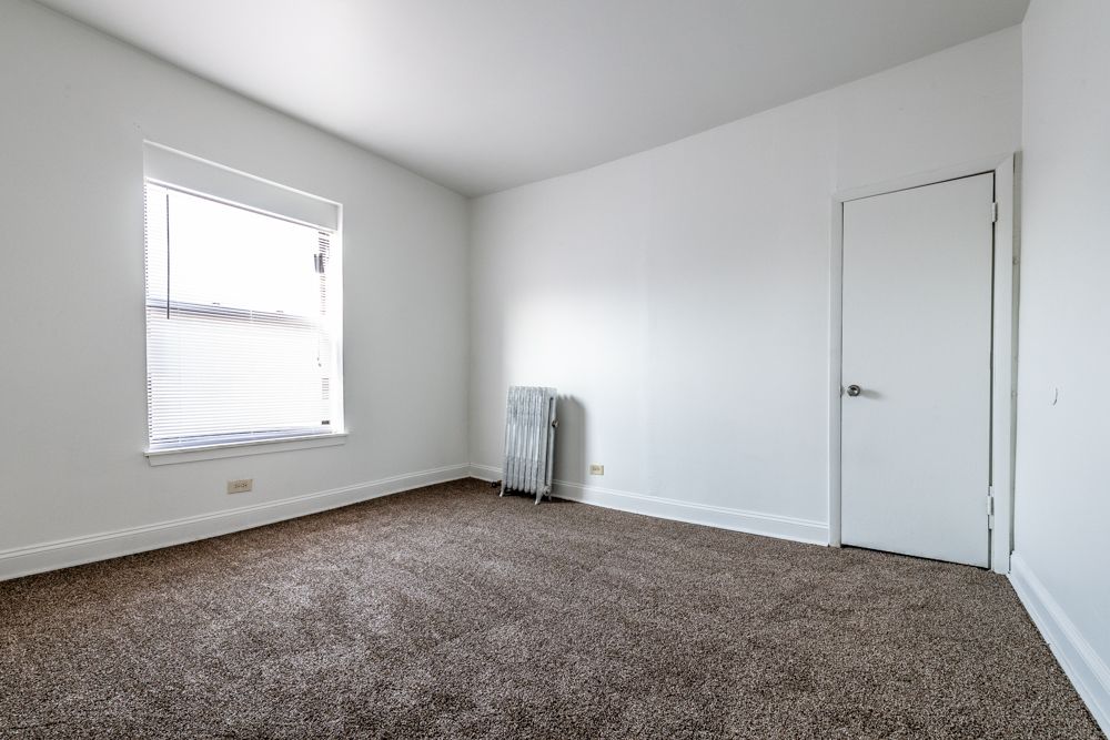 Empty room with white walls, window with blinds, radiator, and carpeted floor.