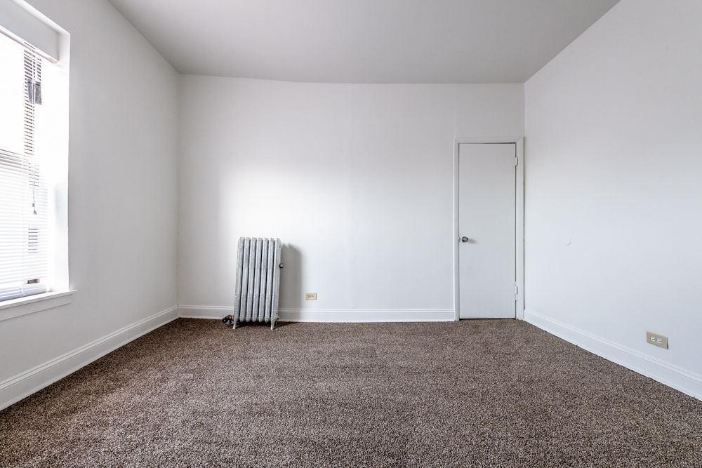 Empty room with brown carpet, white walls, radiator, window, and closed door.