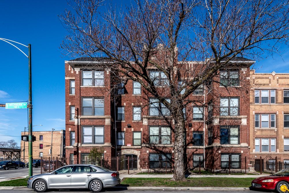 Red brick apartment building with bare tree in front, blue sky. Cars parked on street.