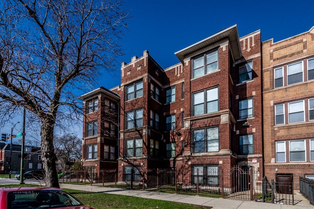Red brick multi-story apartment building with dark windows under a blue sky, street, and tree in front.