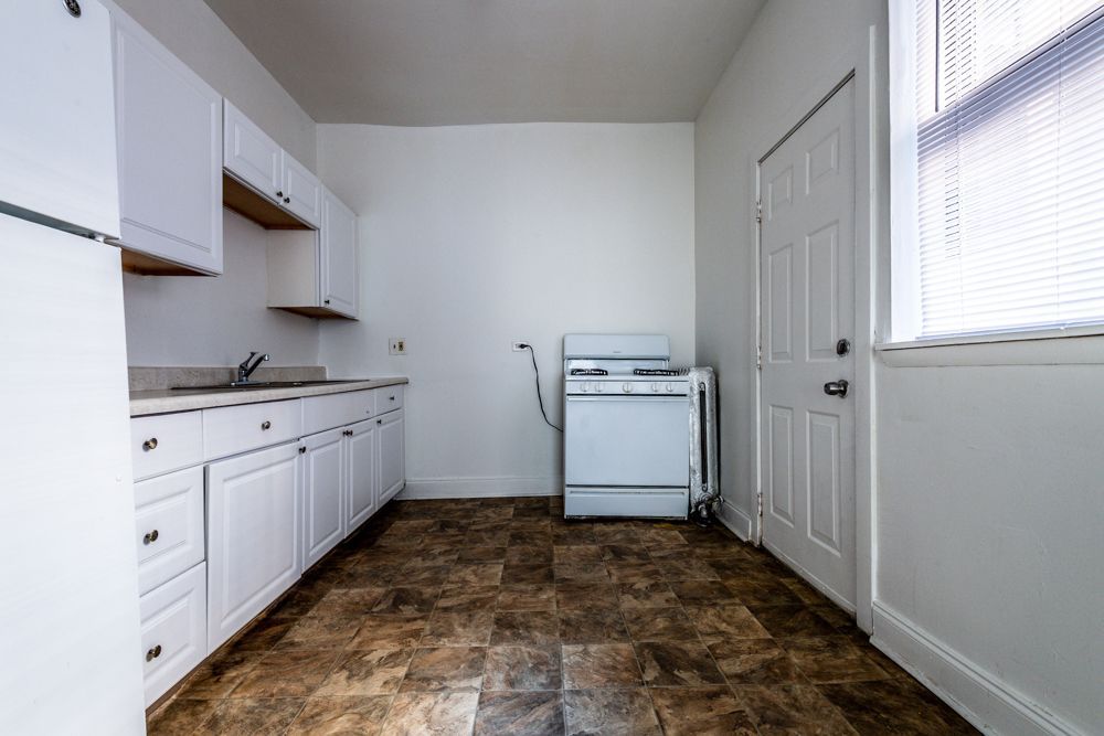 White kitchen with cabinets, stove, sink, and door. Brown tile floor. Blinds on window.