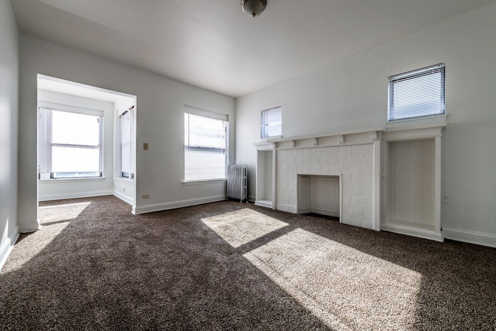Empty room with brown carpet, white walls, small window, radiator.