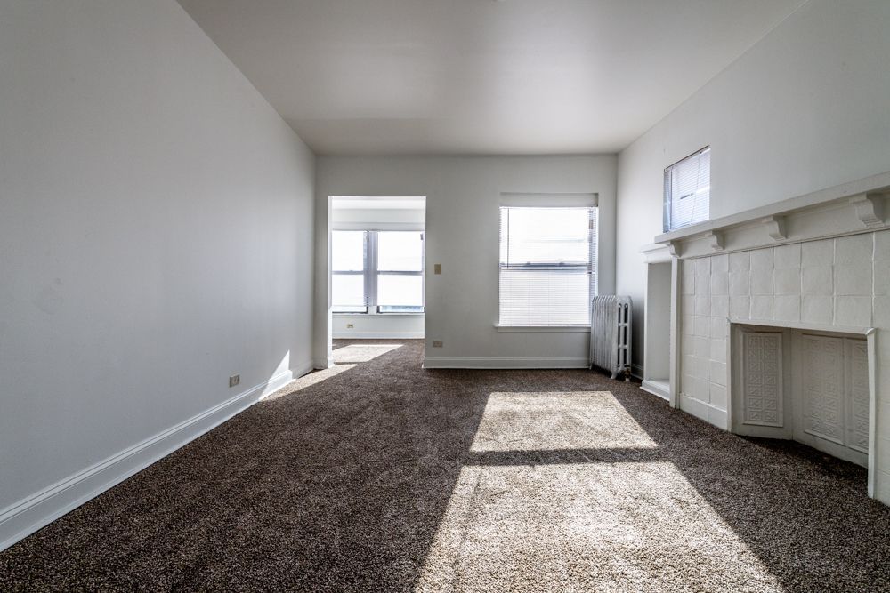 Empty living room with brown carpet, white walls, fireplace, and sunlight through windows.