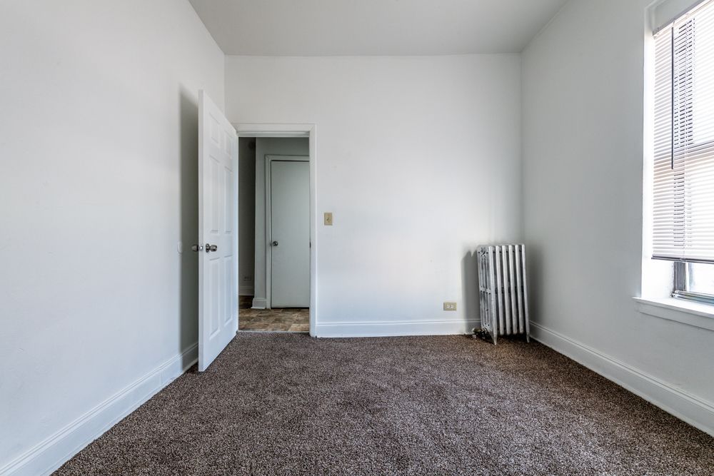 Empty bedroom with brown carpet, white walls, doorway, and radiator near window.