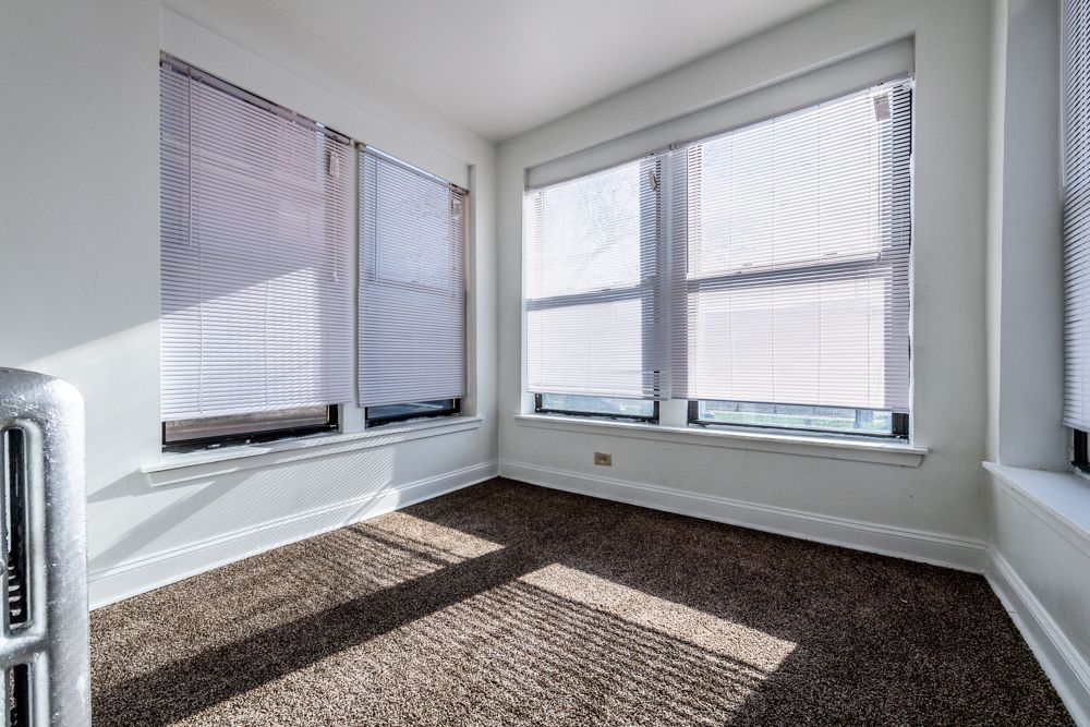 Empty room with three windows, white walls, brown carpet, and blinds. Sunlight streams through.