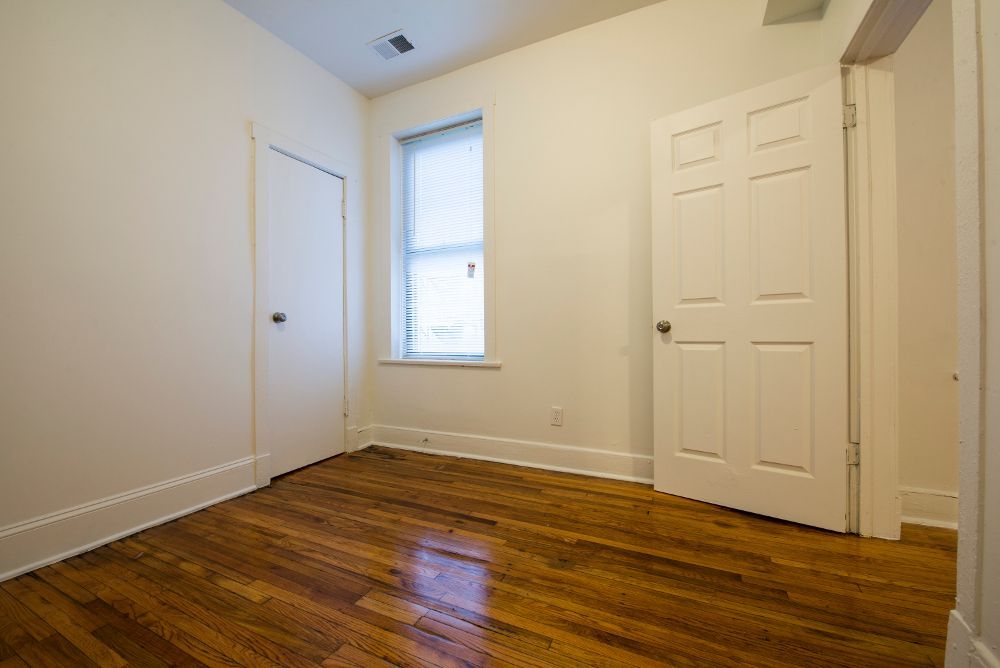 Empty room with hardwood floors, white walls, two doors, and a window with blinds.