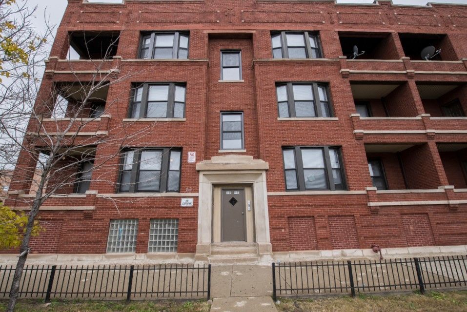 Red brick apartment building with dark-framed windows, a central doorway, and small iron fence.