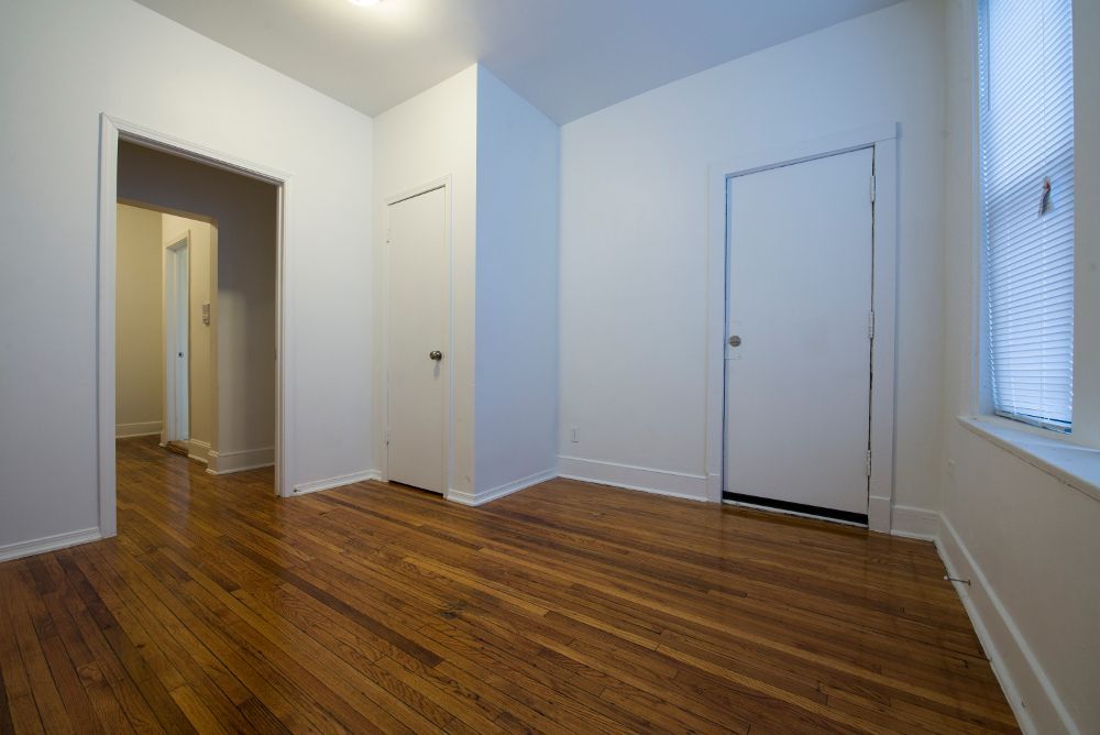 Empty room with hardwood floors, white walls, two doors, and a window with blinds.