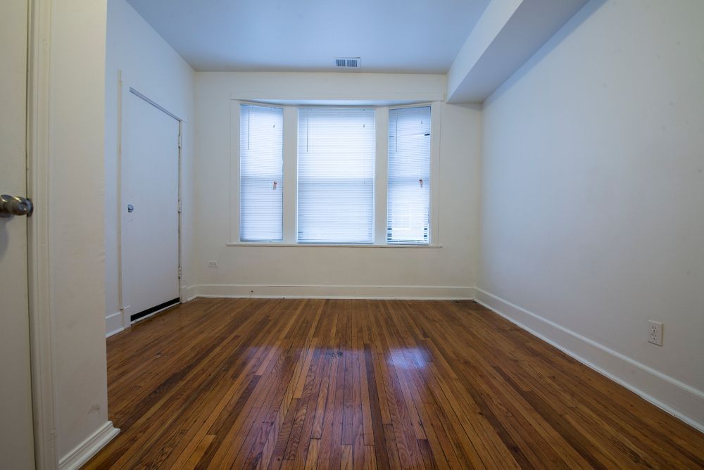 Empty room with hardwood floors, a window with blinds, and a closed door. White walls.