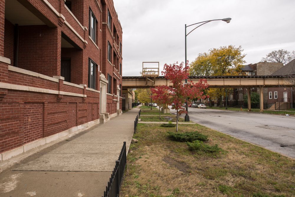 A brick apartment building next to a sidewalk with a train track overhead on a cloudy day.