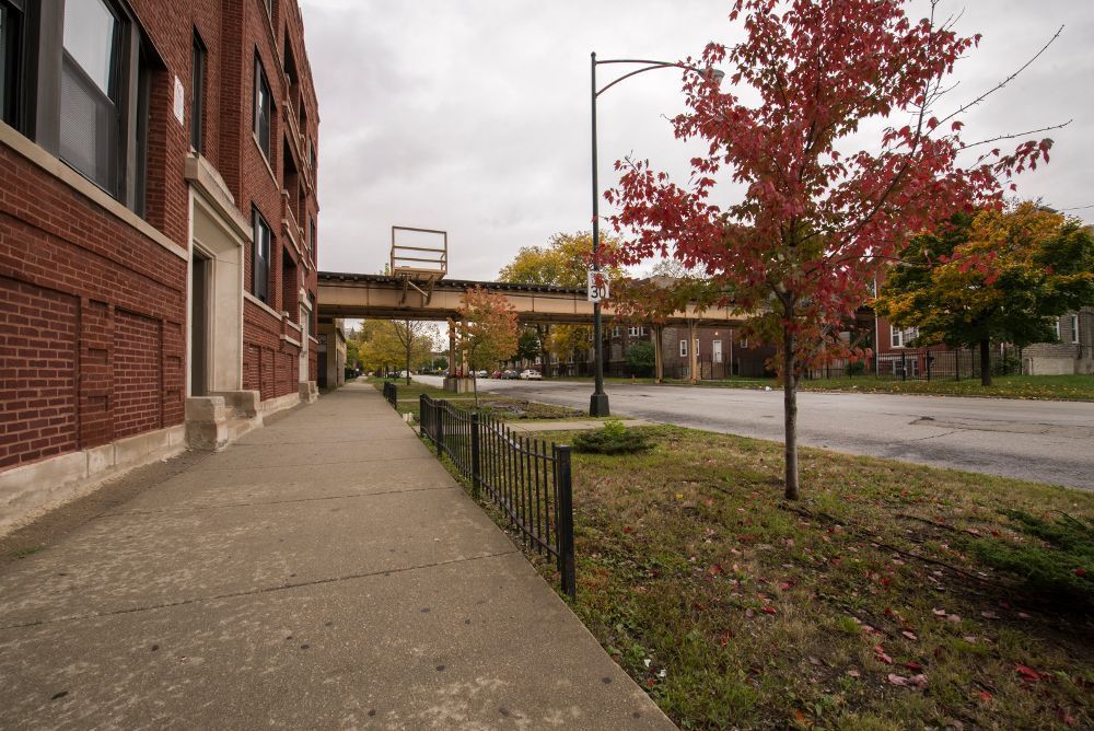Brick building next to a sidewalk with a train overpass in the distance; fall foliage.