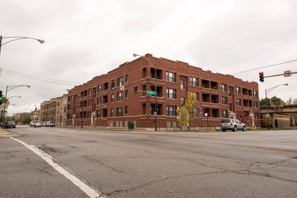 Brick apartment building on a street corner under cloudy sky. Street, cars, traffic lights are visible.