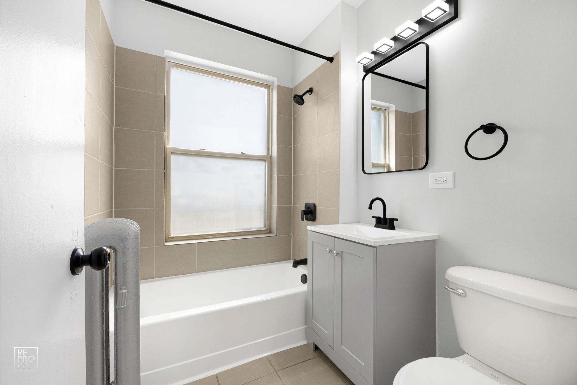Bathroom with light gray walls, white bathtub, vanity, and toilet, with black fixtures and tan tile.
