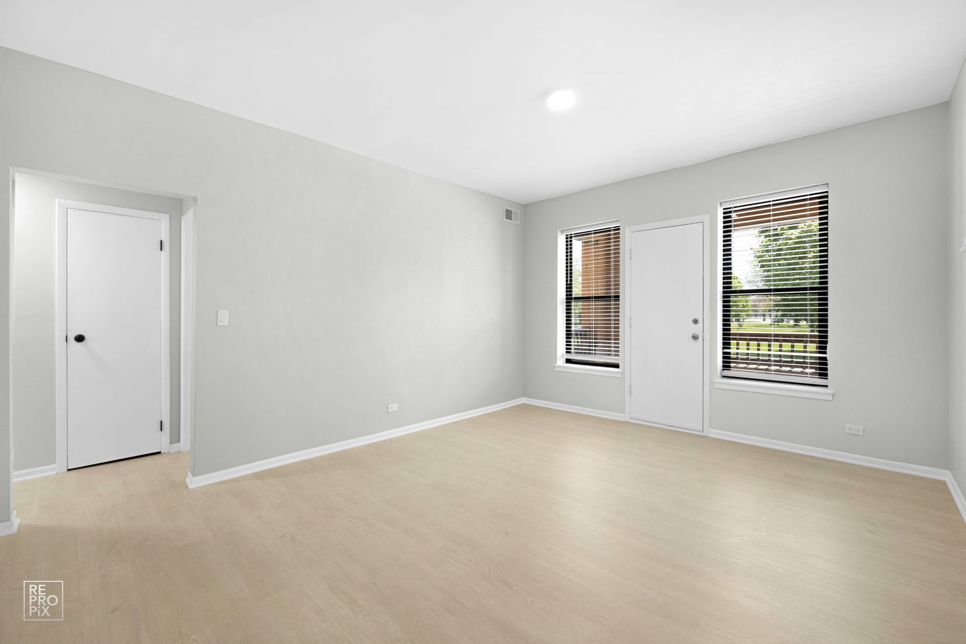 Empty room with light wood floors, gray walls, white door, and windows overlooking greenery.