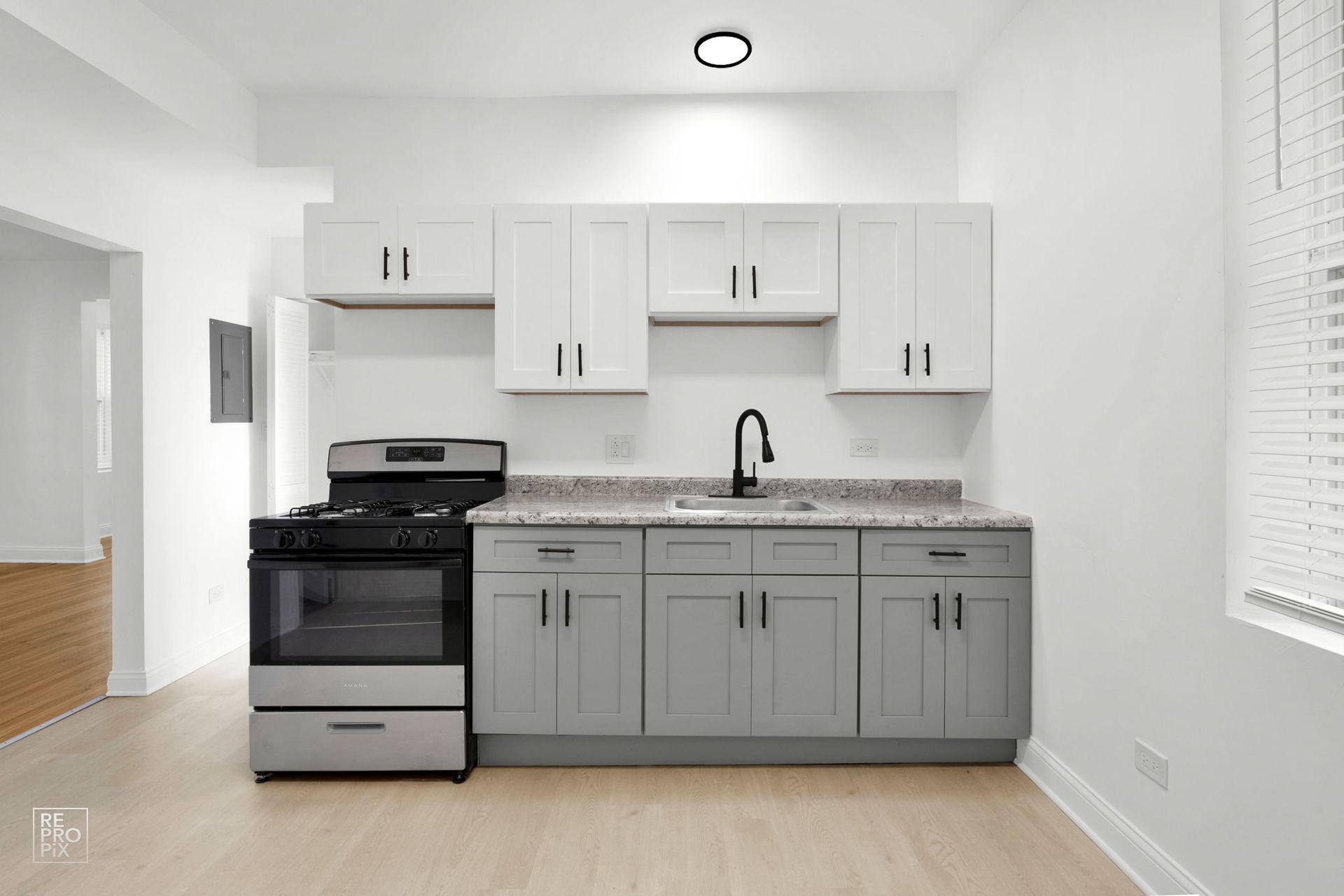 A kitchen with gray and white cabinets, black faucet, stove, and countertops.  Room has wood floors and a window with blinds.