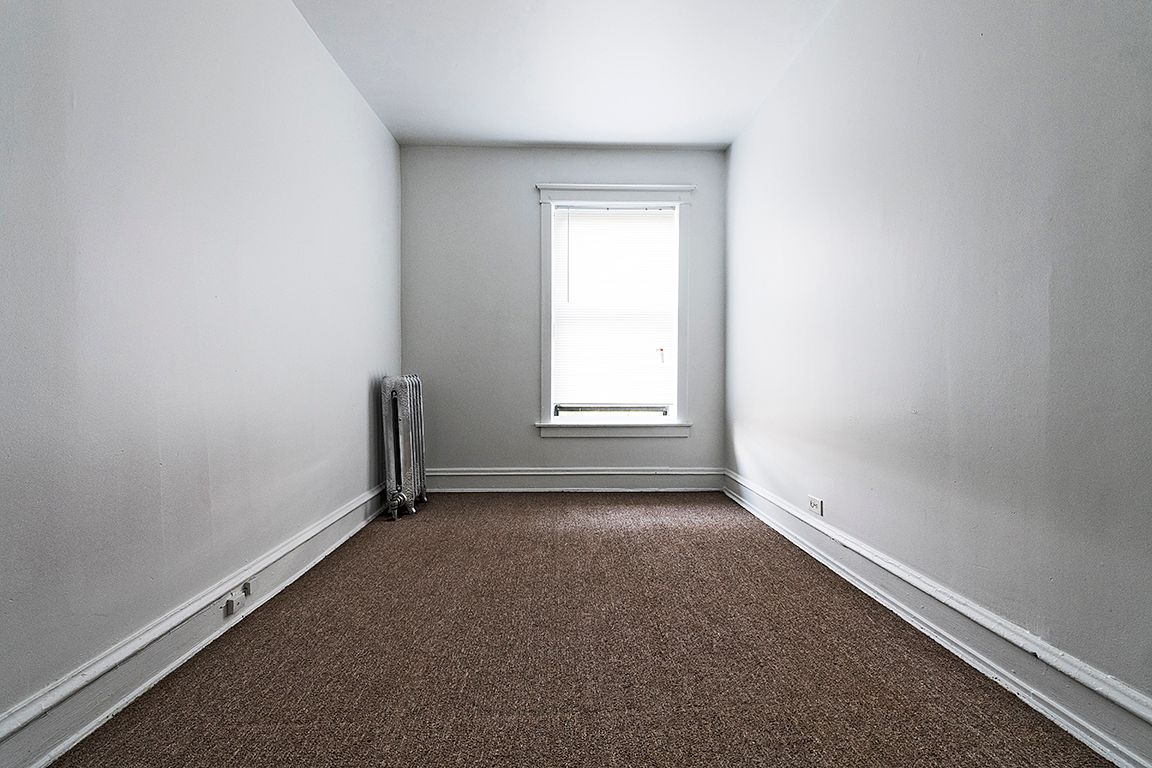 Empty room with brown carpet, radiator on the wall, and window centered at the end.