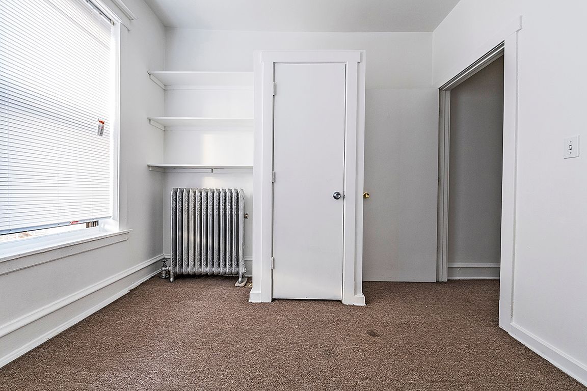 Empty room with brown carpet, white walls, closet door, and shelves. Includes a window and radiator.