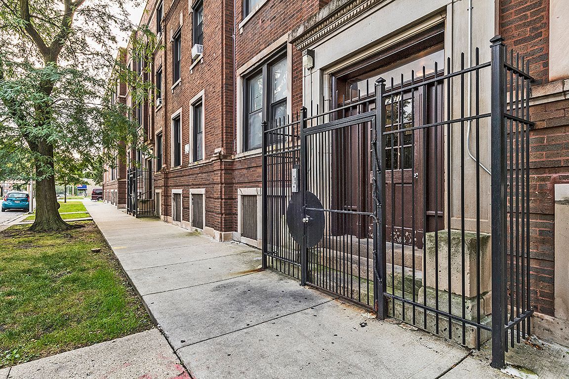 Brick building with a wrought-iron gate and a sidewalk. Trees line the street.