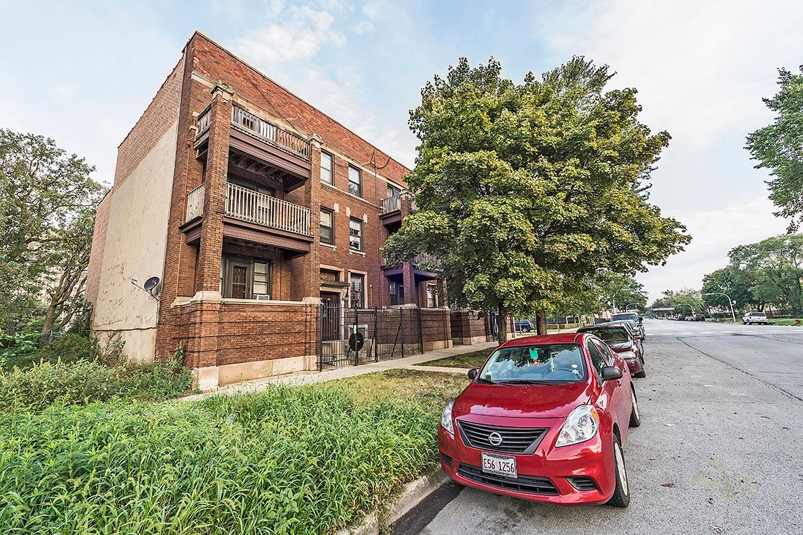 Red brick apartment building with balconies, parked cars on street.