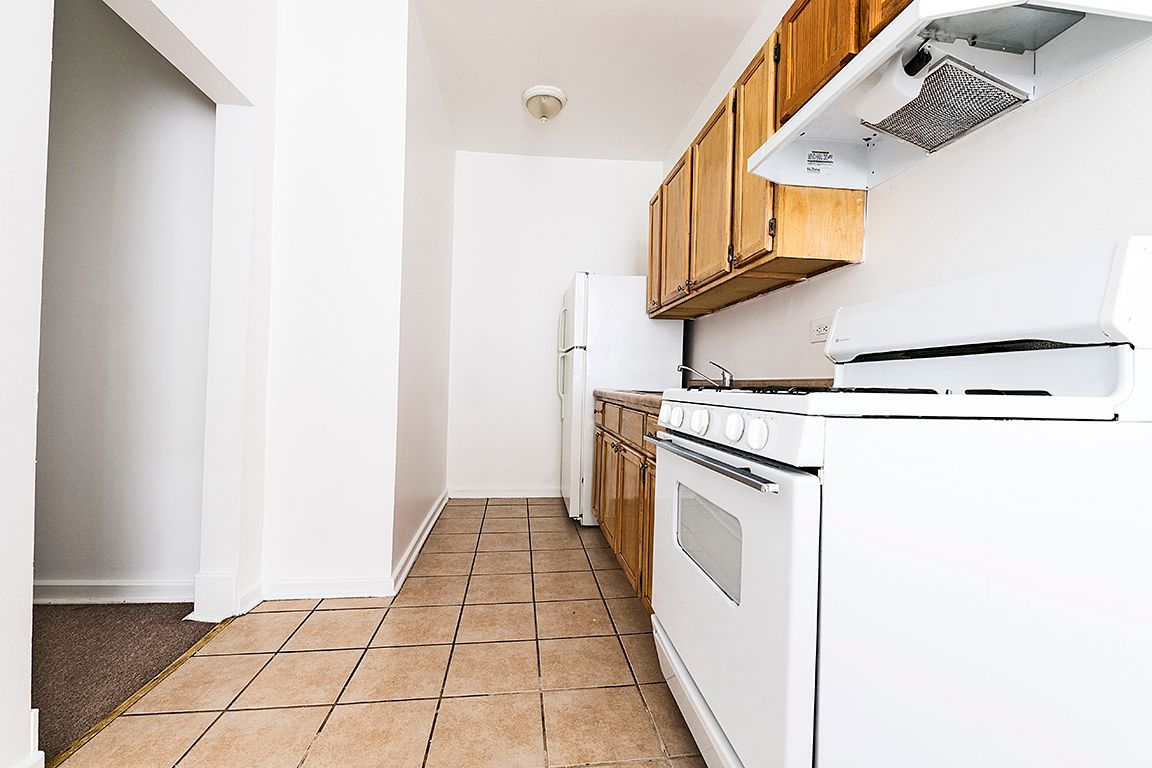 Empty kitchen with white appliances, wooden cabinets, and beige tile floor.